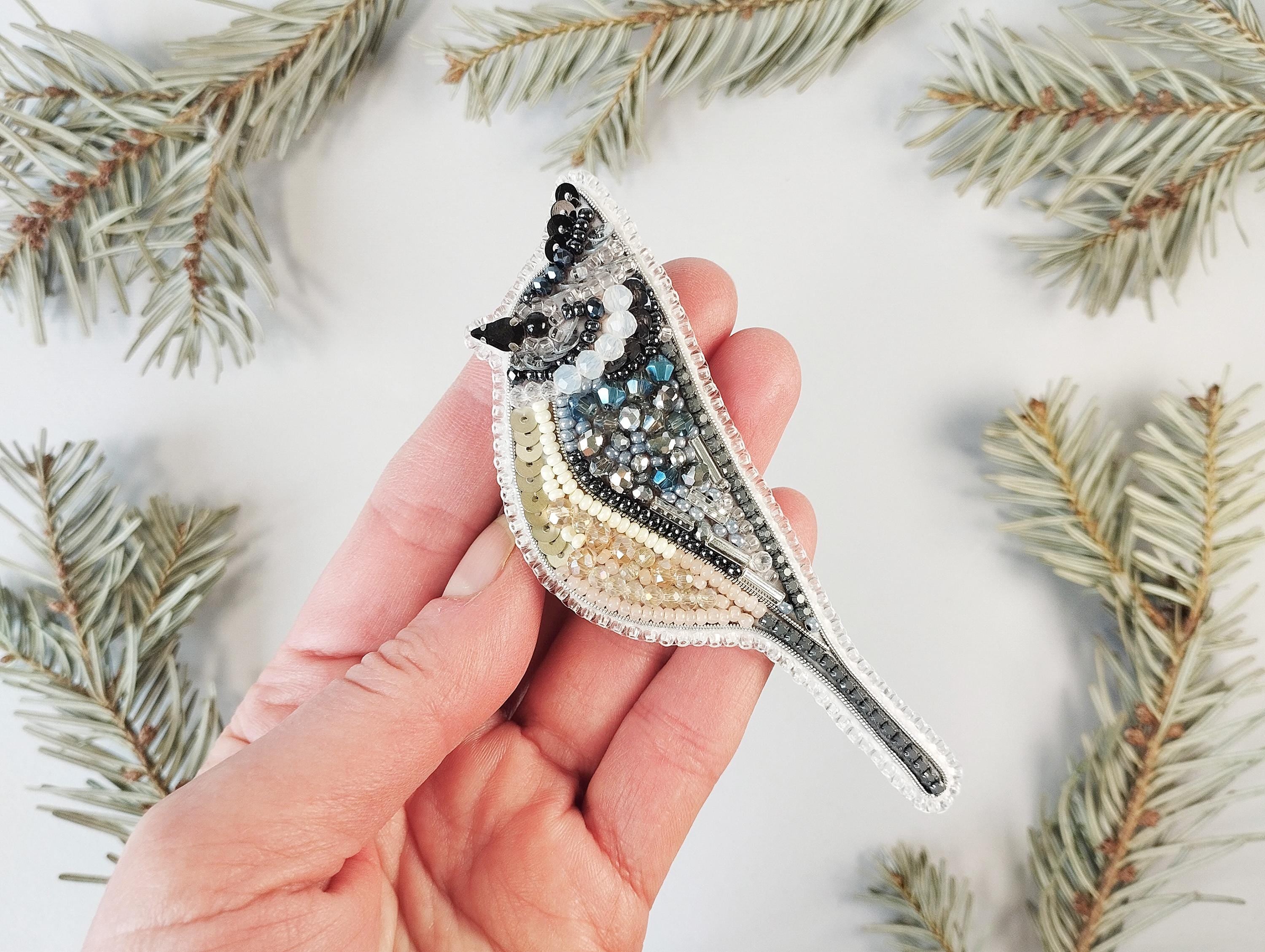 a hand holding a bird brooch with a pine branch in the background
